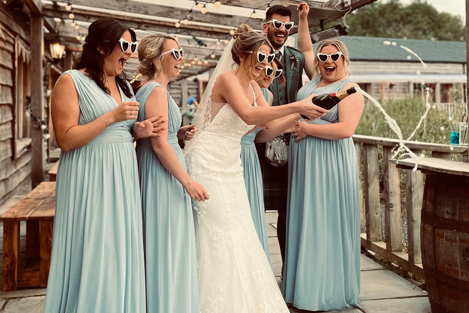 Bride, groom and bridesmaids wearing love heart shaped sunglasses pop bottle of champagne